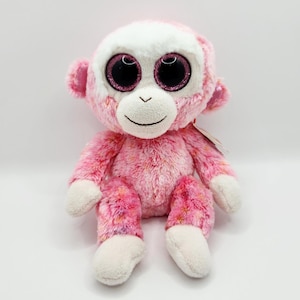 Könnte beinhalten: Ein rosa Plüschaffe mit weißem Gesicht und Füßen. Der Affe hat große, glitzernde Augen und ein lächelndes Gesicht. Das Fell ist eine Mischung aus Rosa und Weiß mit einigen bunten Sprenkeln. Ein Etikett ist am Affen befestigt.