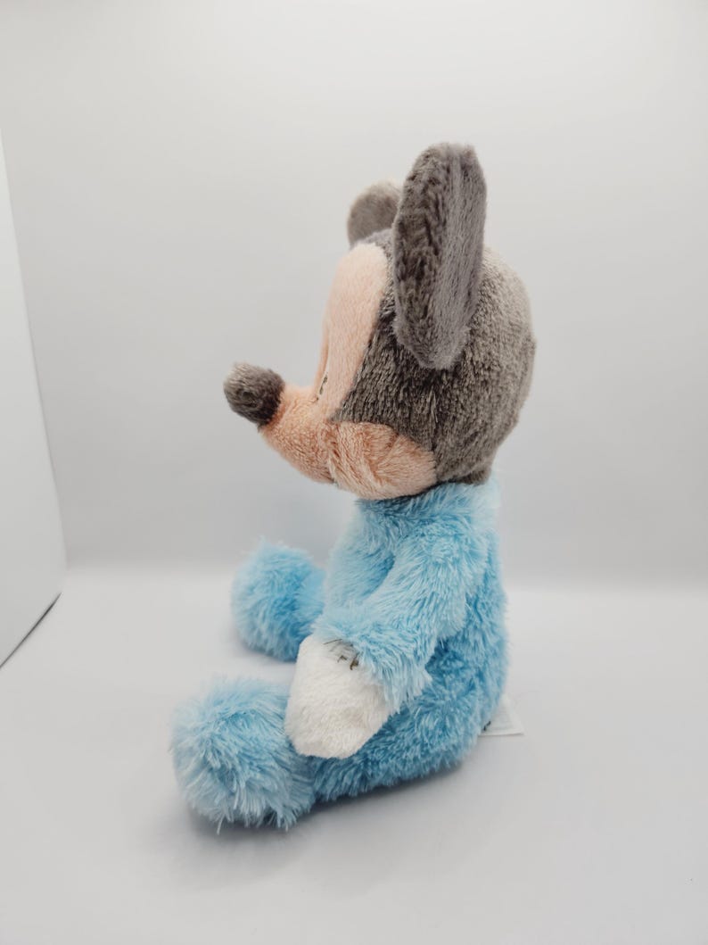 Peut inclure: Une peluche repr&eacute;sentant un personnage de souris, avec une t&ecirc;te et des oreilles grises, un museau rose et un corps bleu clair. Le jouet est assis, avec des d&eacute;tails blancs sur les mains et les pieds. Le jouet est con&ccedil;u pour ressembler &agrave; un personnage de dessin anim&eacute;.