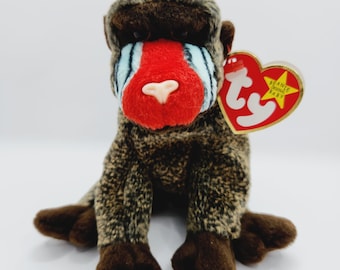 Puede incluir: Un mono de peluche marrón y negro Beanie Baby con la cara roja y rayas azules. El mono está sentado con las piernas cruzadas. El juguete tiene una etiqueta roja en forma de corazón con las palabras "Beanie Babies" y "Baby" en ella.