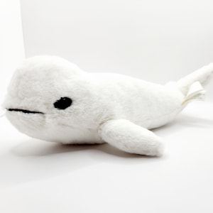 May include: A plush white beluga whale toy with black eyes and a stitched mouth. The soft toy has flippers and a tail, and is designed to resemble a real beluga whale. The toy is made of a soft, fuzzy material.