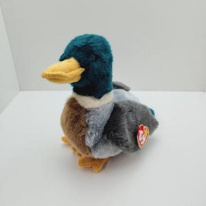 May include: A green, gray, and brown plush duck with a yellow beak and a red Ty tag. The duck is sitting on a white surface.