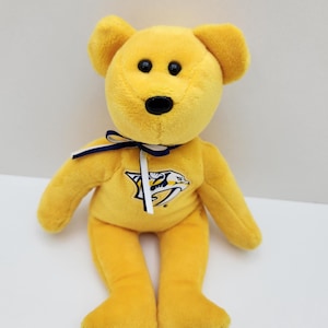 May include: A bright yellow plush bear featuring the Nashville Predators hockey team logo on its chest. The bear has black plastic eyes and a black nose, and it is wearing a thin ribbon tied in a bow around its neck with navy blue and white stripes.