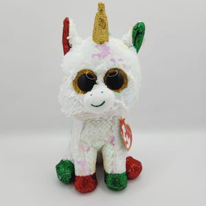 May include: White unicorn plush toy with a gold horn, green and red accents, and large, glittery eyes. The toy is made of sequins and has a Ty tag attached.