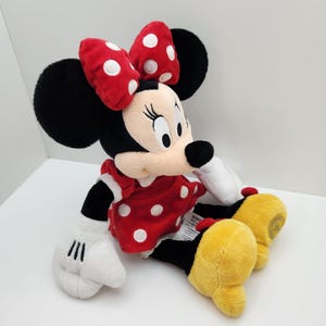 May include: A plush Minnie Mouse toy featuring a red dress with white polka dots, a matching red bow with white polka dots, black ears, white gloves, and large yellow shoes. The character has a light beige face with black eyes and a black nose.