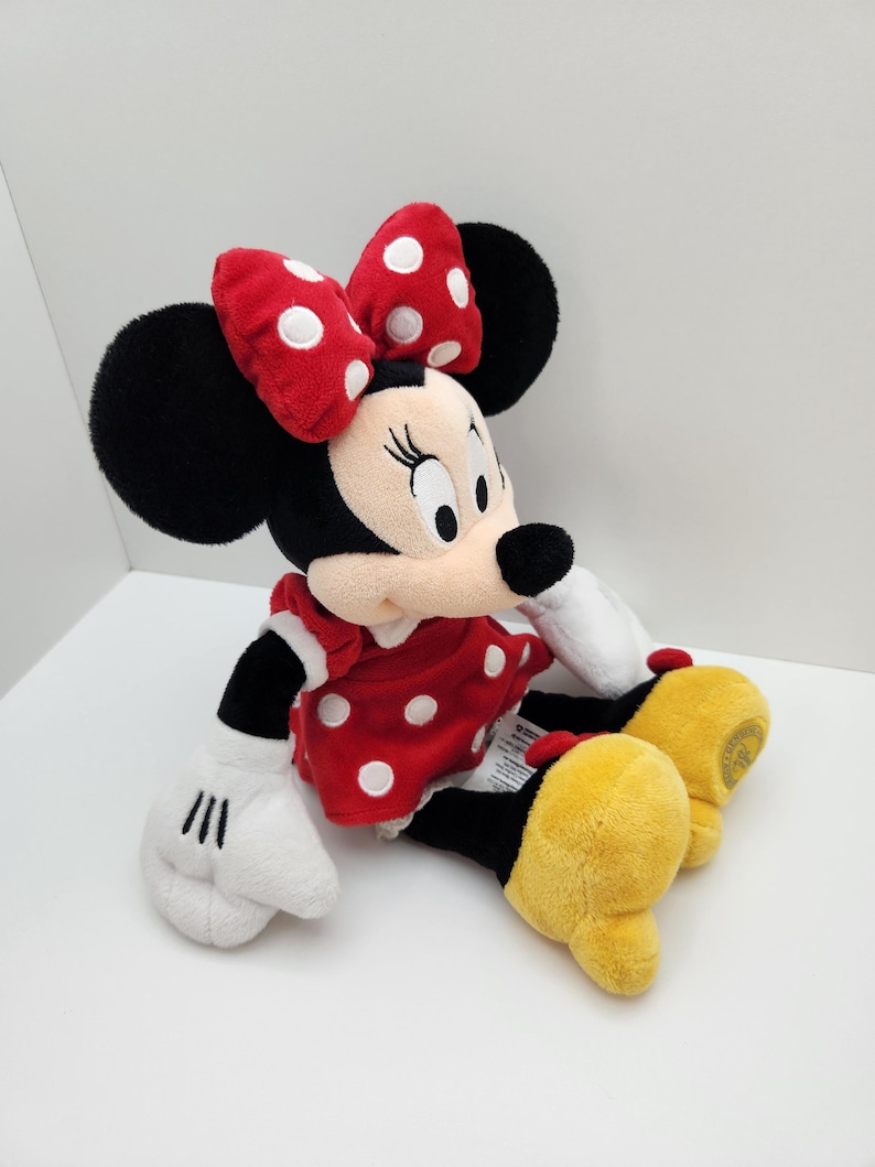 May include: A plush Minnie Mouse toy featuring a red dress with white polka dots, a matching red bow with white polka dots, black ears, white gloves, and large yellow shoes. The character has a light beige face with black eyes and a black nose.