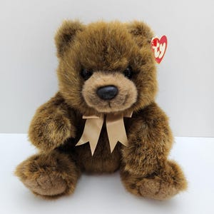 May include: A plush teddy bear with shaggy brown fur, dark eyes, and a black nose. The bear features a light tan satin ribbon tied in a bow around its neck and a red heart-shaped Ty tag attached to its ear. The toy is seated in an upright position.