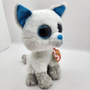 May include: A plush toy wolf with white and gray fur, blue accents on the ears and nose, and large, glittery black eyes. The toy has a red heart-shaped tag attached.