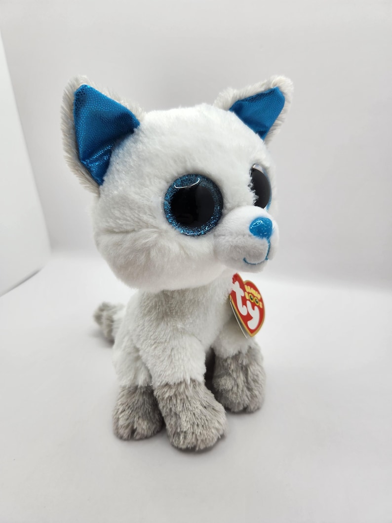 May include: A plush toy wolf with white and gray fur, blue accents on the ears and nose, and large, glittery black eyes. The toy has a red heart-shaped tag attached.
