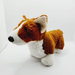 May include: A brown and white plush toy dog with floppy ears. The dog is sitting on a white surface.