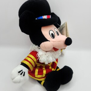 Op de afbeelding: Een Mickey Mouse pluche speelgoed gekleed in een rood en goud militair jasje en een zwarte hoed. De hoed heeft een blauw, wit en rood lint. Mickey's gezicht is roze met zwarte ogen en een zwarte neus. Het speelgoed heeft witte handschoenen en zwarte schoenen.