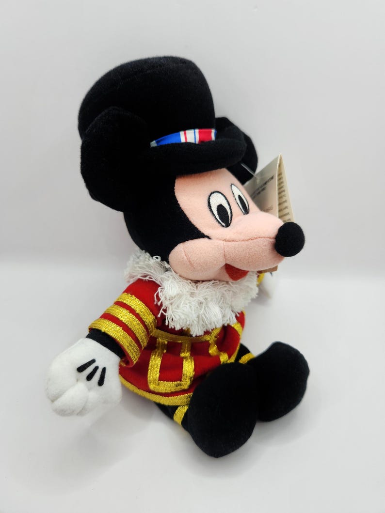 Op de afbeelding: Een Mickey Mouse pluche speelgoed gekleed in een rood en goud militair jasje en een zwarte hoed. De hoed heeft een blauw, wit en rood lint. Mickey's gezicht is roze met zwarte ogen en een zwarte neus. Het speelgoed heeft witte handschoenen en zwarte schoenen.