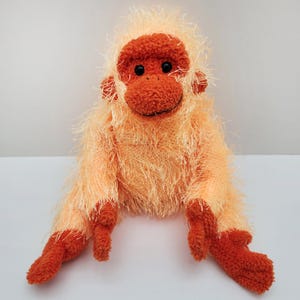 May include: A soft toy monkey in shades of orange. The monkey has a fuzzy body and a round face with black eyes and a smiling mouth. Its arms and legs are extended. The toy is made from a soft, plush material, perfect for children.