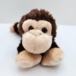 May include: A plush monkey toy with brown fur and a tan face, hands, and feet. The monkey is lying down, with its arms and legs extended. The toy has black eyes and a friendly expression.