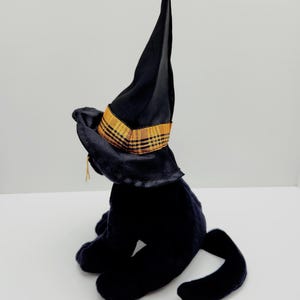 Puede incluir: Un gato de peluche negro con un sombrero de bruja negro con una banda a cuadros dorada y naranja. El gato est&aacute; sentado con la cola enroscada. El sombrero tiene una corona alta y puntiaguda y un ala ancha. Decoraci&oacute;n de Halloween.