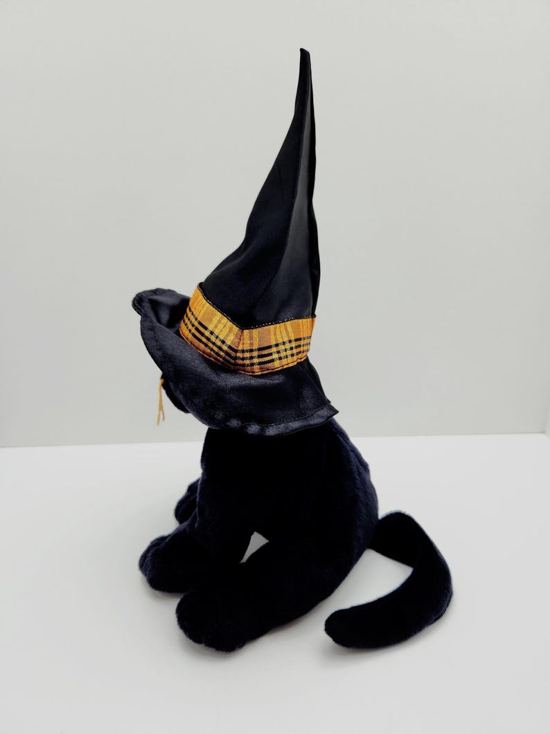 Puede incluir: Un gato de peluche negro con un sombrero de bruja negro con una banda a cuadros dorada y naranja. El gato est&aacute; sentado con la cola enroscada. El sombrero tiene una corona alta y puntiaguda y un ala ancha. Decoraci&oacute;n de Halloween.