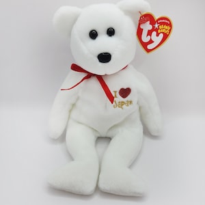 May include: White plush beanie baby bear with a red ribbon around its neck and a red heart with the words "I Love Japan" embroidered on its chest. The bear has black eyes and a black nose. The bear is sitting on a white background. The bear has a Ty tag attached to its ear that says "Original Beanie Babies".