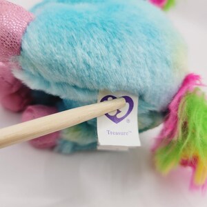 Op de afbeelding: Een blauwe en groene pluche eenhoorn met een roze manen en staart. De eenhoorn heeft een paarse tag met de tekst "Treasure" met een paars hartlogo.