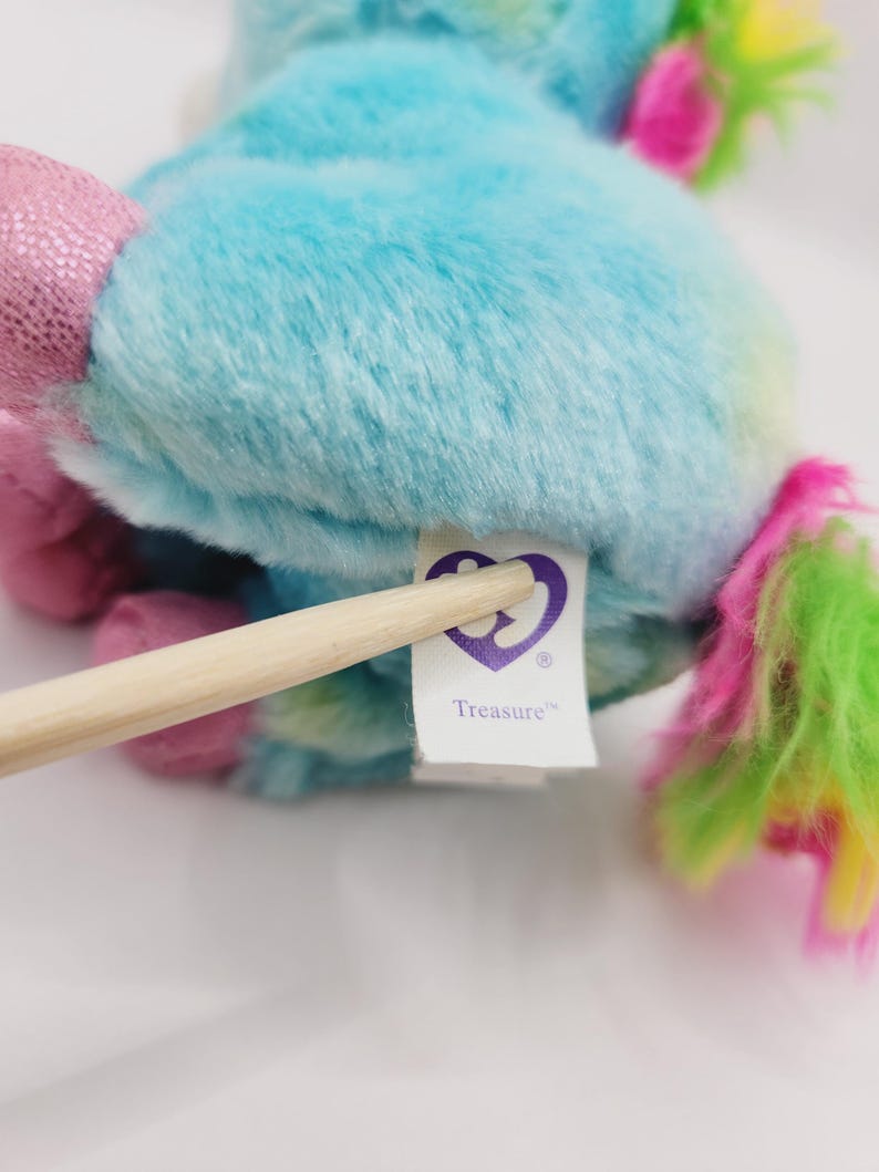 Op de afbeelding: Een blauwe en groene pluche eenhoorn met een roze manen en staart. De eenhoorn heeft een paarse tag met de tekst "Treasure" met een paars hartlogo.