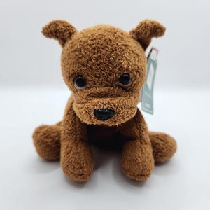 May include: A brown plush dog toy with large, dark eyes and a black nose. The stuffed animal is sitting with its paws forward and has a tag attached. The toy is made of a soft, textured material.