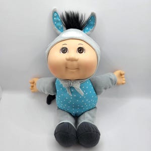 Peut inclure: Poupée Cabbage Patch Kids vêtue d'un costume de petit âne gris à pois bleus. La poupée a les yeux bruns et un sourire. Le costume comprend une capuche avec des oreilles bleues et une petite queue noire.