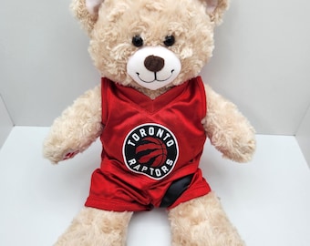 May include: A plush teddy bear with light tan fur, black eyes, and a brown nose. The bear is wearing a red basketball jersey and matching red shorts. The jersey features the Toronto Raptors logo, which includes a black basketball graphic.