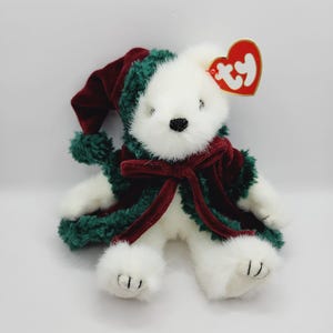 May include: White plush teddy bear wearing a green and red robe with a red velvet hat. The bear has a Ty tag attached to its ear.