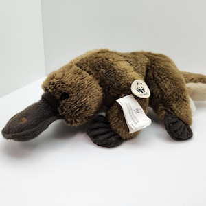 May include: A plush platypus toy with brown fur, a dark brown bill, and a flat tail. The toy has a tag with the WWF logo. The toy is lying on a white surface.