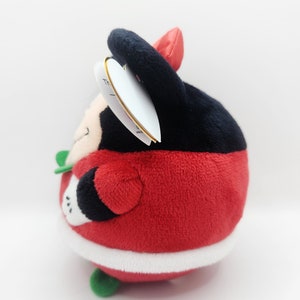 Puede incluir: Juguete de peluche rojo y negro con forma de personaje redondo que lleva un gorro de Pap&aacute; Noel. El juguete tiene un ribete blanco alrededor de la parte inferior y detalles verdes.