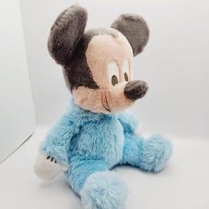 Peut inclure: Une peluche de Mickey Mouse en position assise. Le jouet est v&ecirc;tu d'une barboteuse bleu clair, avec un visage beige, un nez noir et des oreilles grises. Des accents blancs sont visibles sur les mains et les pieds.