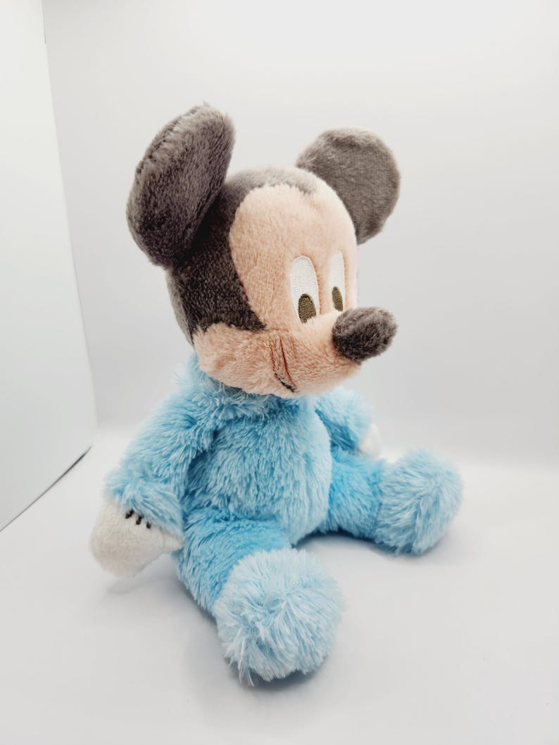 Peut inclure: Une peluche de Mickey Mouse en position assise. Le jouet est v&ecirc;tu d'une barboteuse bleu clair, avec un visage beige, un nez noir et des oreilles grises. Des accents blancs sont visibles sur les mains et les pieds.