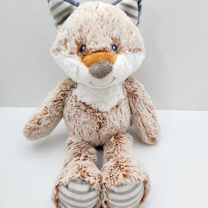 May include: A plush fox toy with a soft, textured body in shades of brown, white, and orange. The fox has blue and gray striped ears and paws. The toy is designed to sit upright, with its arms and legs extended.