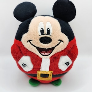 May include: A red and white plush Mickey Mouse toy wearing a Santa Claus costume. The toy is round and has a smiling face with black eyes and a black nose.