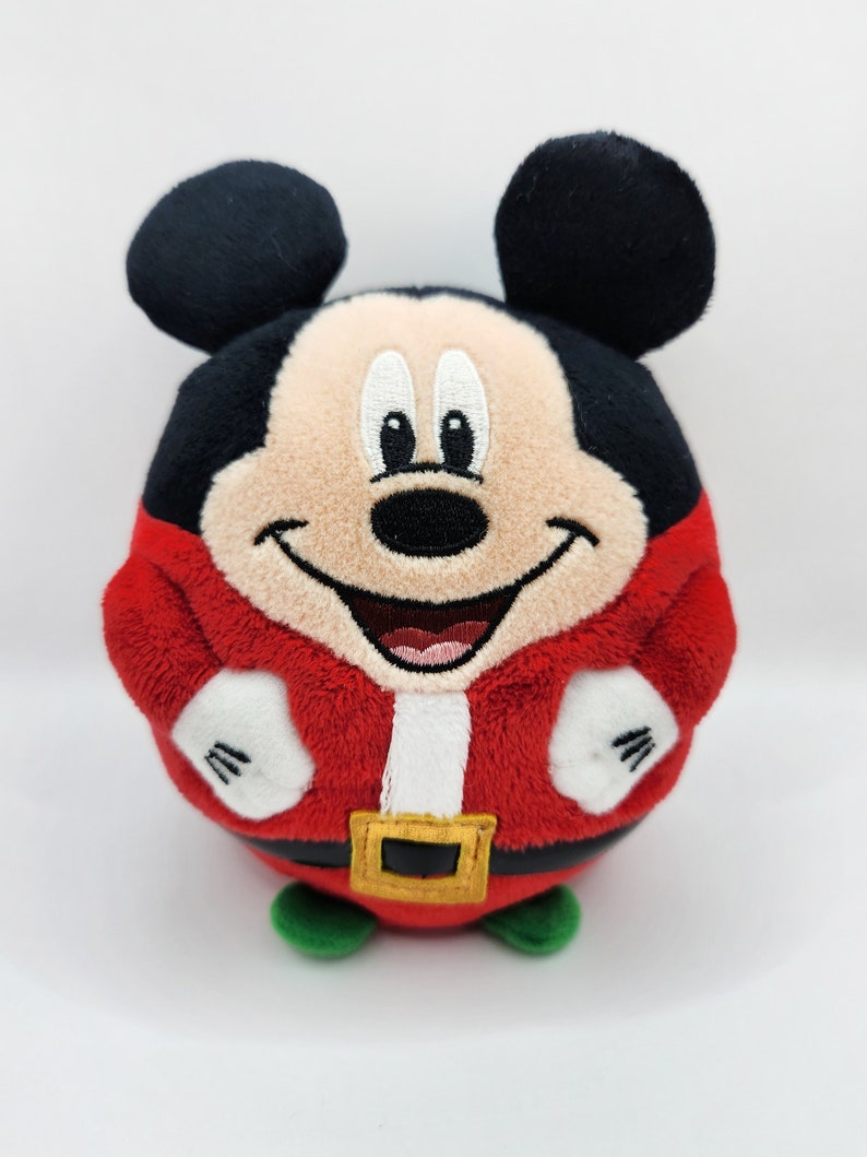 May include: A red and white plush Mickey Mouse toy wearing a Santa Claus costume. The toy is round and has a smiling face with black eyes and a black nose.