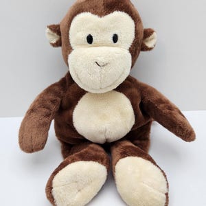 May include: A plush monkey toy featuring soft brown fur on the head, arms, and back, contrasted with a cream-coloured face, ears, chest, and feet. The monkey has small black embroidered eyes and a simple stitched mouth, sitting in an upright position.