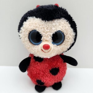 Può includere: Un peluche a forma di coccinella con un corpo rosso soffice a pois neri e un viso color crema. Presenta grandi occhi rotondi blu scuro, un piccolo naso rosso e due piccole antenne rosse sulla testa nera e soffice. Ha braccia e gambe nere.