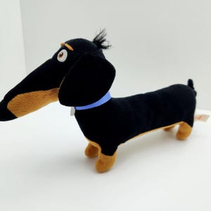 Può includere: Un peluche a forma di cane bassotto nero con dettagli marroni e un collare blu. Il cane ha un lungo muso e orecchie flosce. Giocattolo dal design stravagante, probabilmente un personaggio cinematografico.
