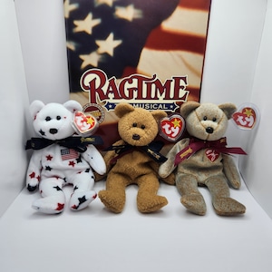 Könnte beinhalten: Drei Ty Beanie Baby Plüschtiere, ein weißer Bär mit roten und blauen Sternen, ein brauner Bär und ein brauner Bär. Die Bären tragen rot-weiß-blaue Bänder und haben Ty-Etiketten angebracht. Die Bären sitzen vor einem Buch mit dem Titel "Ragtime Musical".