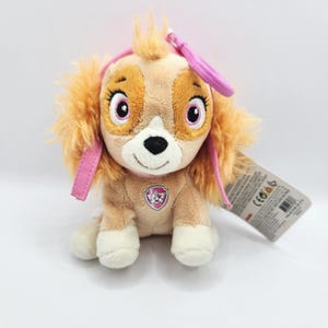 May include: A plush toy of a cartoon dog with tan fur, pink accents, and a pink clip. The dog has large, expressive eyes and a smiling face. A tag is attached to the toy.