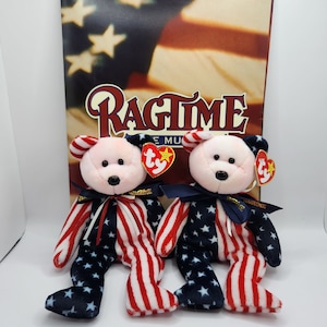 Può includere: Due orsacchiotti Beanie Baby rosa e bianchi che indossano abiti patriottici rosso, bianco e blu. Gli orsacchiotti sono seduti davanti a una rivista con la parola "Ragtime" sulla copertina.
