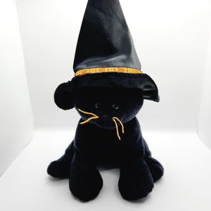 Puede incluir: Un juguete de peluche negro con forma de gato que lleva un sombrero de bruja negro con una banda dorada. El gato tiene bigotes de hilo amarillo y est&aacute; sentado sobre sus cuartos traseros. El juguete est&aacute; sobre un fondo blanco.