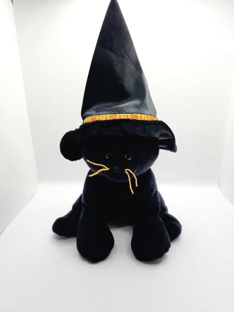 Puede incluir: Un juguete de peluche negro con forma de gato que lleva un sombrero de bruja negro con una banda dorada. El gato tiene bigotes de hilo amarillo y est&aacute; sentado sobre sus cuartos traseros. El juguete est&aacute; sobre un fondo blanco.