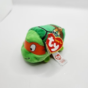 May include: A green plush toy of a Teenage Mutant Ninja Turtle character, with an orange mask and a brown shell design. The toy has a red and white Ty tag attached. The toy is a Teeny Ty.