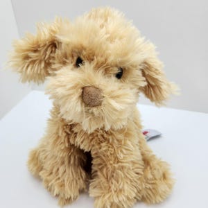 May include: A plush stuffed dog toy with shaggy, light tan fur, dark brown plastic eyes, and a soft, textured brown nose. The toy is in a seated position with floppy ears and a small fabric tag visible on the side of its body.