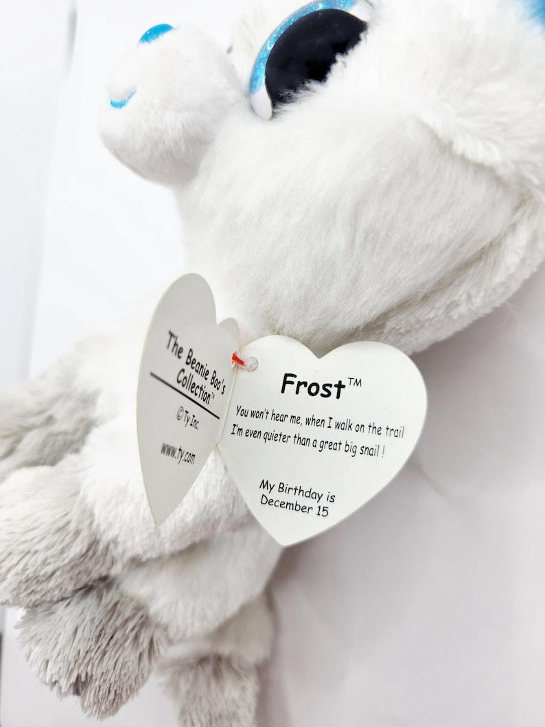 May include: A white plush toy with blue accents and large, glittery eyes. The toy has a heart-shaped tag that reads "Frost" and includes a poem and the birthday, December 15. Part of The Beanie Boo's Collection.