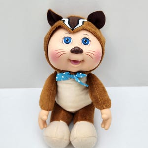 May include: A plush doll in a brown squirrel costume with a light tan bodysuit. The doll has blue eyes, rosy cheeks, and a blue bow tie with white polka dots. The doll's ears are brown.