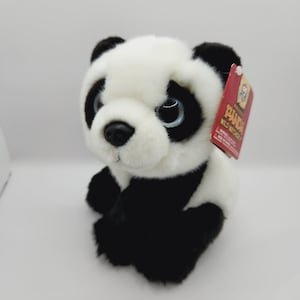 May include: A black and white plush panda bear toy with large eyes. The toy is sitting on a white surface and has a red tag attached to its ear that says "Precious Panda Wild Watchers".
