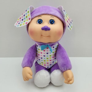 May include: A plush doll wearing a purple bunny costume with a hood and ears. The doll has a light skin tone, rosy cheeks, and blue eyes. The costume features a white bodice with colorful heart patterns and a blue bow tie.