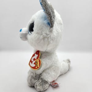 May include: A white plush toy fox with large, sparkly black eyes and gray accents on the ears and paws. The fox has a red tag with gold trim attached to its ear. The toy is sitting on a white surface.