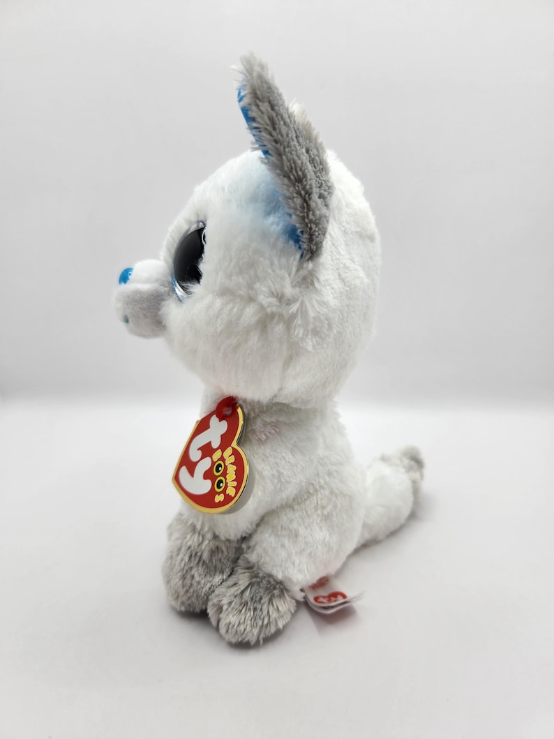 May include: A white plush toy fox with large, sparkly black eyes and gray accents on the ears and paws. The fox has a red tag with gold trim attached to its ear. The toy is sitting on a white surface.
