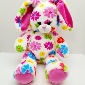 May include: A plush bunny with a white body covered in colorful flower patterns. The bunny has long ears with pink lining, pink paws, and a pink nose. The flowers are in shades of pink, blue, yellow, and green.